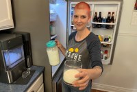 A photo of a woman standing by a refrigerator and holding a pitcher and glass of raw milk.