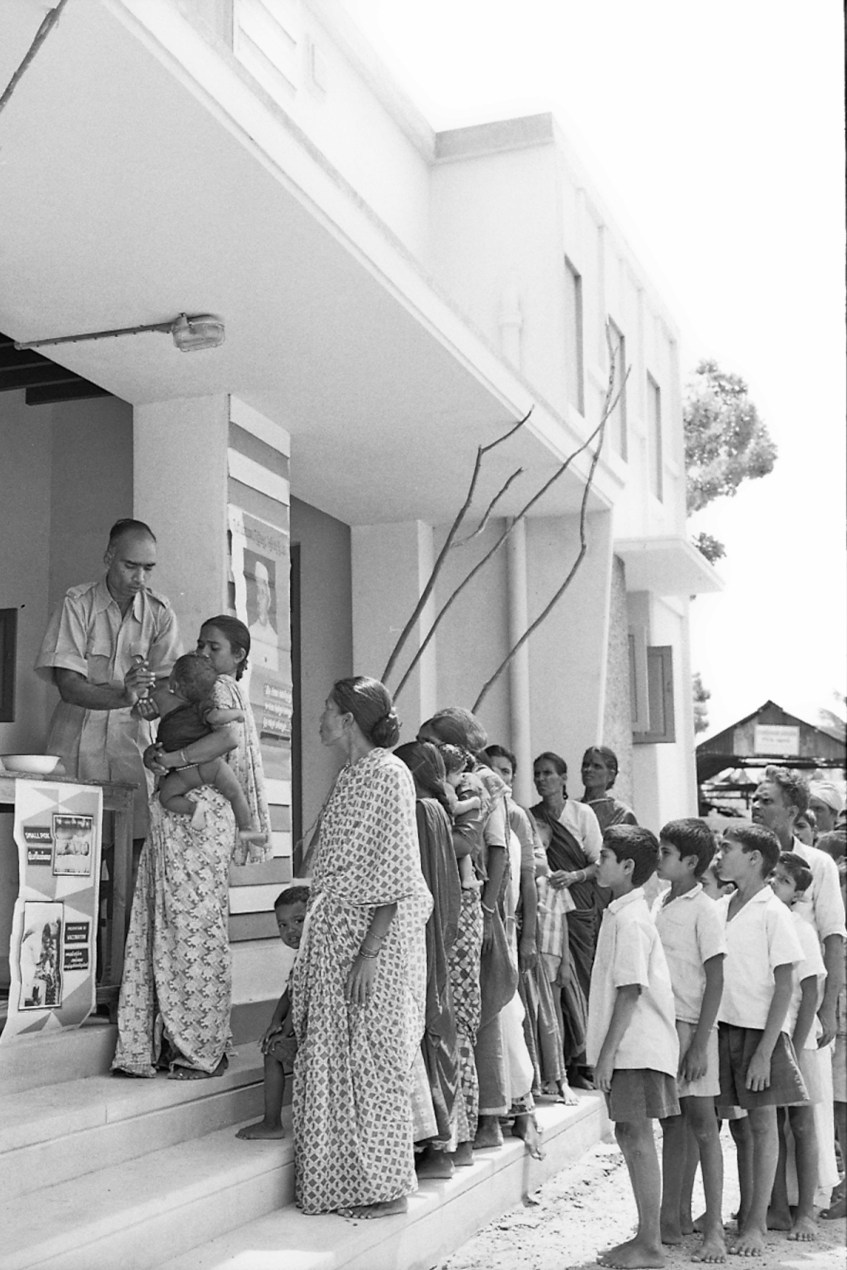 People of all ages and genders line up outside a vaccination station. The photograph is black-and-white, taken by a film camera in 1962.