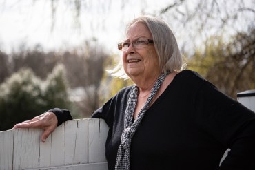 A photo of an older woman standing for a portrait outside by a fence.