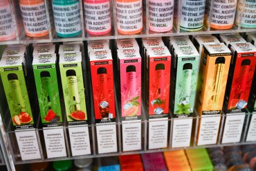A photo of a row of colorful vaping and e-cigarette products labeled with fruit flavors and a warning that they contain nicotine.