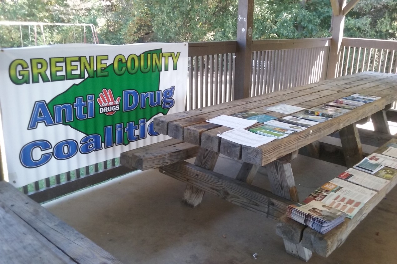 A wooden picnic table has paper sand brochures laid out on the table and a bench. A banner reading "Greene County Anti-Drug Coalition" hangs on a raining behind the table.