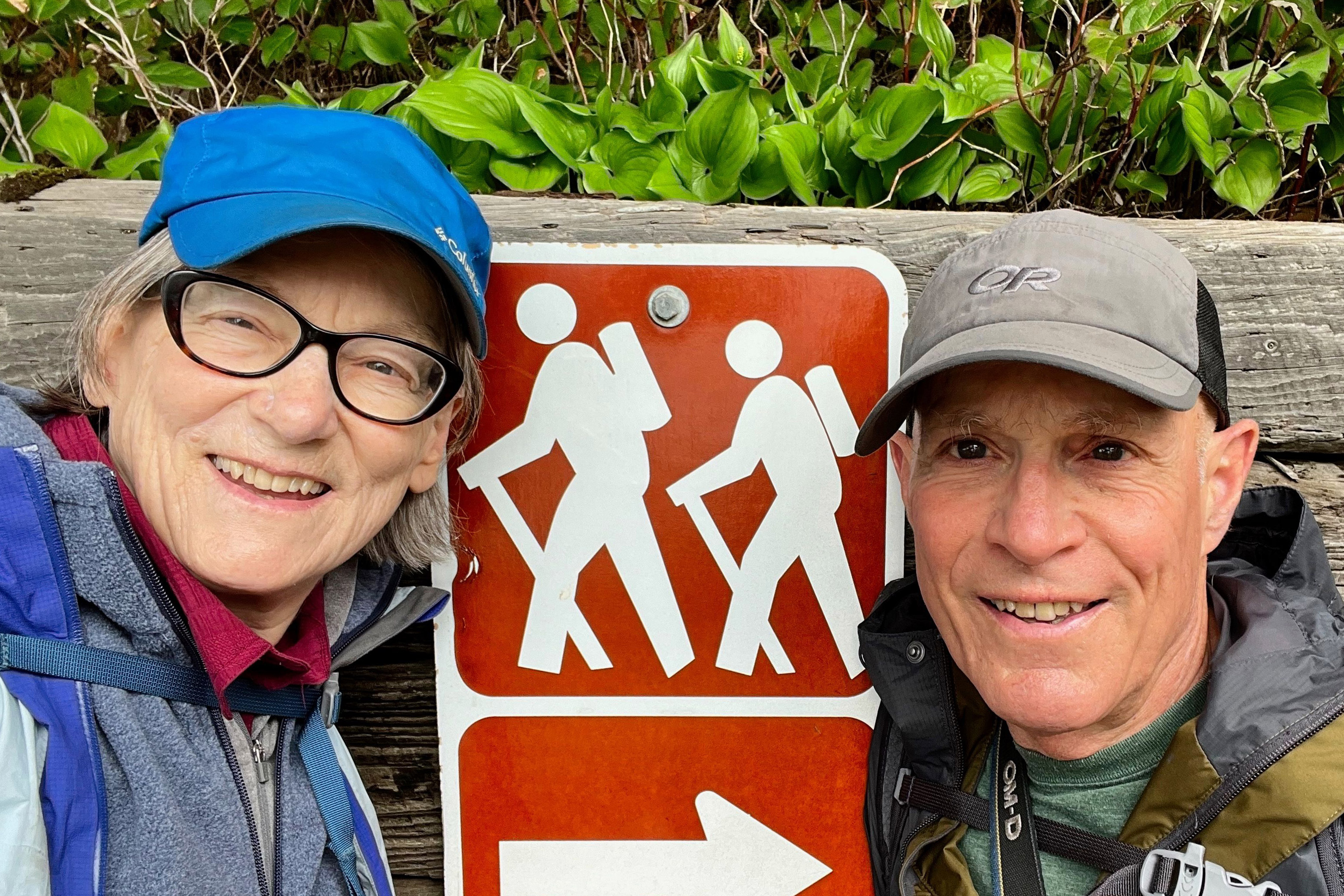 A photo of a couple posing for a selfie while outdoors in front of a hiking trail sign.